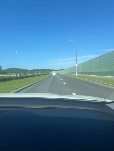 The image shows a view from inside a vehicle traveling on a highway, likely in or near Cherepovets, Russia. The road is asphalt with white lane markings. On the right side of the road, there is a tall, pale green sound barrier. On the left side of the road, there is a grassy embankment lined with hedges, followed by trees and a blue sky. Several streetlights are visible along the roadside, and a few cars are in front of the vehicle, moving in the same direction. The dashboard of the vehicle is visible in the foreground, reflecting the sky and road.