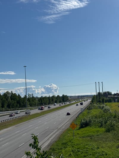 This is a photograph taken from an elevated vantage point, looking down upon a multi-lane highway in Anchorage, Alaska, United States. The sky is a vibrant blue with scattered white clouds. The highway, appearing to be a section of the Seward Highway, is filled with a moderate amount of traffic. Several vehicles, including cars and a red truck, are visible on the roadway. On the right side of the highway, a green grassy embankment is present, with scattered bushes and trees. A yellow diamond-shaped sign indicating "RIGHT LANE ENDS" is visible in the foreground, along with a speed limit sign for 55 mph. On the left side of the highway, there are more trees and some buildings in the distance. Power lines and a tall street light are also visible. The overall impression is a typical highway scene in a suburban or semi-urban environment on a clear day.