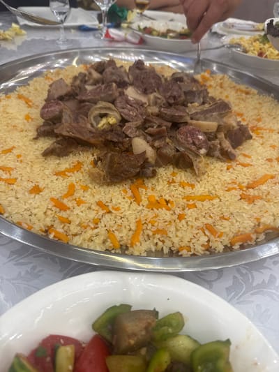 A close-up, eye-level shot captures a large platter of plov, a rice dish, topped with a generous portion of cooked meat and sliced sausage, arranged on a silver tray placed on a white tablecloth. Interspersed within the fluffy rice are shredded orange carrots. In the foreground, a white bowl holds a colorful salad composed of diced tomatoes, cucumbers, and green bell peppers. The scene appears to be set in a restaurant in Jezqazğan, Kazakhstan, where a hand is visible in the upper right corner, using a fork to lift a portion of the plov. In the background, glassware and other dining elements suggest a communal meal.