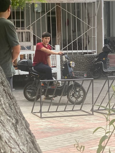 In Kokand City, Uzbekistan, a young male is seated on a black electric scooter. He is wearing a red t-shirt and dark trousers. The scooter has a storage compartment on the back and a basket in the front. The scooter is parked on a paved surface with red and white checkered tiles. In the background, there is a building with a white fence and a stone wall. Another black scooter is parked behind the first one. A person wearing a green shirt is standing to the left of the frame, facing away from the camera. A large tree trunk is visible in the foreground on the left side of the image.