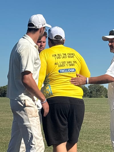 Four men are gathered on a grassy field in Port Alfred, South Africa, under a clear blue sky. The central figure, viewed from the back, wears a bright yellow long-sleeved shirt featuring the text "FOR ALL THE CRIMES I DID LAST NIGHT ALL I GOT WAS THIS LOUSY T-SHIRT" and a "TRELLIDOR" logo. He is also wearing black shorts and a white baseball cap. To his immediate left, a man in a white polo shirt, soiled light-colored long pants, and a white baseball cap stands with his left side visible; a blue and white wristband is on his left wrist. Between these two, a third man's face is partially visible. To the right, a fourth man, wearing a white shirt and a white baseball cap, has his left hand placed on the central figure's back, near the waistline, and also wears a blue and white wristband on his left wrist. Trees are visible in the distant background.