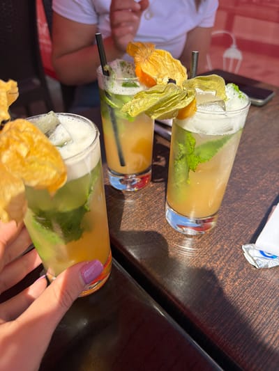 The image shows three cocktails on a dark wooden table, likely outdoors. The cocktails are tall and cylindrical, with a golden-yellow liquid and green mint leaves visible inside. Each cocktail is garnished with a physalis fruit, its papery husk partially open to reveal the orange berry within. One cocktail is being held by a hand with a purple fingernail. A person in a white shirt is partially visible in the background. The edge of a mobile phone is also visible on the table. The location is most likely not in the city of Guildford, United Kingdom as the cocktail presentation is not typical. There is no evidence that the photo was taken in that location.