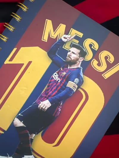 A close-up, slightly angled shot captures a spiral-bound notebook featuring a prominent image of Lionel Messi in his Barcelona uniform. The notebook's cover is predominantly blue and burgundy, with large yellow numbers and lettering that read "10 MESSI". Messi is depicted in mid-action, with his right arm raised and index finger pointing upwards, suggesting a celebration or emphasis. He is wearing a blue and red striped Barcelona jersey with his team's crest visible on the chest, and a captain's armband on his left arm. His black shorts and red-striped socks are also visible, along with a Nike logo on his shorts. The notebook is propped up, and the background appears to be a red fabric, possibly a tablecloth or clothing. The image's quality suggests it was taken with a mobile phone. There is no visible indication of Villa de Cura, Venezuela within the image itself.