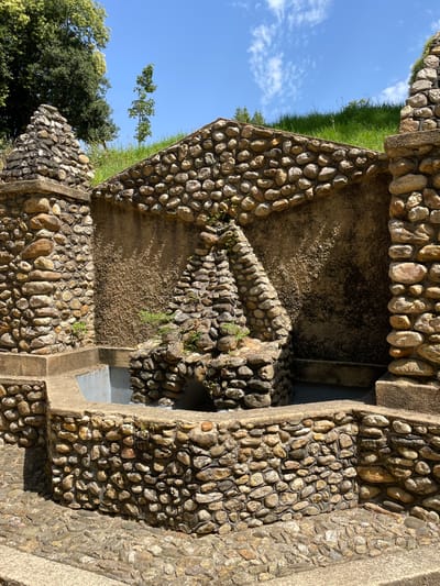 The image depicts an outdoor stone fountain in a park-like setting, likely in Águeda, Portugal, given the context. The fountain is constructed primarily from natural stones of varying sizes and colors, with some areas featuring rough, textured plaster. The main structure of the fountain is semicircular, with a lower basin area. Rising behind the basin is a tiered, pyramid-like stone structure that appears to be the focal point of the fountain, though it is not actively spouting water in the image. Small green plants and moss are growing in crevices of the stone structure. The fountain is surrounded by a paved area made of smaller, irregularly shaped stones. In the background, a grassy incline rises, topped with lush green trees and a bright blue sky with scattered white clouds. The sun casts shadows on the fountain and the surrounding area, indicating a bright, sunny day. There are no people visible in the image.
