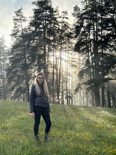 In Yundola, Bulgaria, a young woman with long blonde hair and glasses stands in a grassy field dotted with yellow wildflowers. She is wearing a dark quilted jacket and black leggings. The background features a dense forest of tall pine trees, with sunlight filtering through the branches and creating a misty atmosphere. The ground is covered in lush green grass and the small yellow flowers. The scene is set outdoors, under a cloudy sky.