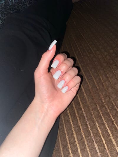 A close-up, high-angle shot shows a left hand with white, square-shaped artificial nails resting on a dark brown textured surface. The hand is positioned from the wrist upwards, with fingers slightly spread and bent. The skin appears pale with some redness around the cuticles. The background consists of a dark, possibly black, fabric on the left and the brown textured surface on the right. There is a hint of a patterned fabric in the top left corner. The lighting casts shadows from the fingers onto the brown surface. This image was taken in the city of Pesnevo, Russia.