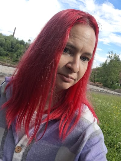 A medium shot selfie taken outdoors shows a woman with bright red hair, facing slightly to her right. She has light-colored eyes and her mouth is slightly open. She is wearing a light-colored shirt layered under a plaid jacket with stripes of purple, gray, and white. A wooden button is visible on the jacket. The background is a grassy field with some small white and yellow flowers. In the distance, there are trees, a cloudy sky, and a tall, thin industrial structure or smokestack. The scene appears to be located on the outskirts of Sim, Russia, possibly in an industrial or construction area with surrounding natural elements.