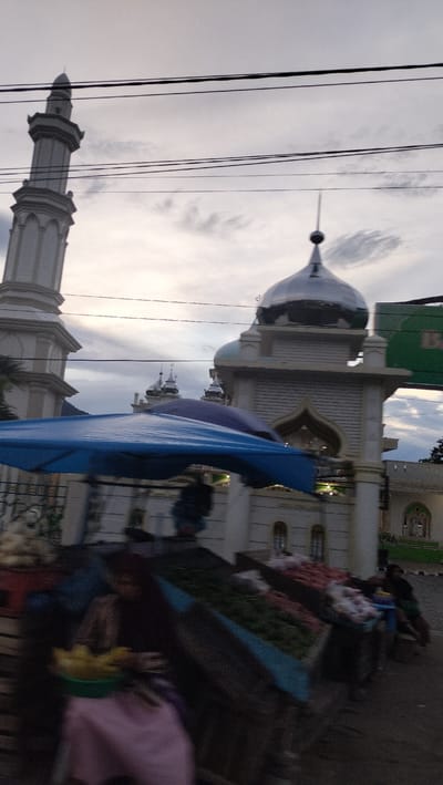This image captures a street scene in Sukamulia, Indonesia, featuring a mosque and what appears to be a local market. The photograph seems to be taken from a moving vehicle or person, as indicated by the motion blur, especially in the foreground and mid-ground.

In the background, a prominent mosque stands out against a cloudy, overcast sky. On the left, a tall, multi-tiered white minaret with classical Islamic architectural elements reaches towards the sky. To the right, the main mosque structure is visible, characterized by a large, reflective silver dome and several smaller domes. The mosque building itself is light-colored, possibly white or cream, featuring decorative archways and facade details. Several power lines cut across the upper portion of the sky, passing in front of the mosque.

In the mid-ground and foreground, elements of a market are present. A bright blue umbrella or canopy partially covers market stalls, under which produce like yellow fruits in a green bowl and various vegetables (reddish and green items) are displayed on what appear to be wooden carts or stands. Several human figures are present around these stalls, though they are blurred and indistinct due to motion and distance. One figure is partially visible under the blue canopy, and another person is seated near the market stalls on the right side of the frame.

The overall lighting suggests either an overcast day or the soft light of early morning or late afternoon. The sky is filled with light gray clouds, indicating cloudy weather. The scene depicts a common sight of daily life, where commercial activity takes place adjacent to a significant religious building. No discernible text is clearly visible on any signs or structures within the image.