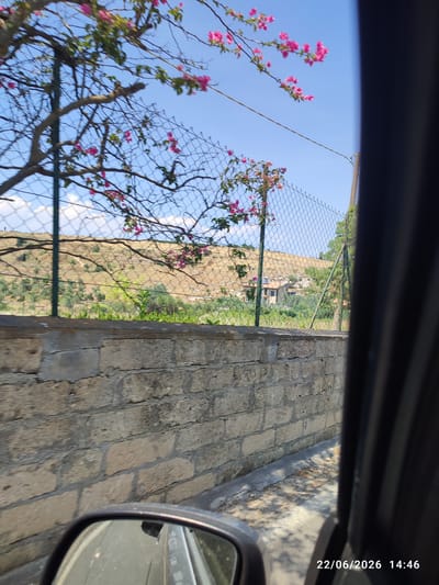This image shows a view from a car in Militello in Val di Catania, Italy. The scene is framed by a rough brick wall in the foreground and a dark car interior on the right. Beyond the wall and fence, a hilly landscape is visible with scattered trees and vegetation. A house with a terracotta roof is nestled on the hillside. Pink bougainvillea flowers bloom on branches that extend over a chain-link fence, adding a splash of color to the bright blue sky dotted with a few white clouds. The time stamp at the bottom right of the image indicates it was taken on 22/06/2026 at 14:46, suggesting it is a sunny afternoon. The overall impression is one of a picturesque rural or semi-rural Italian setting.