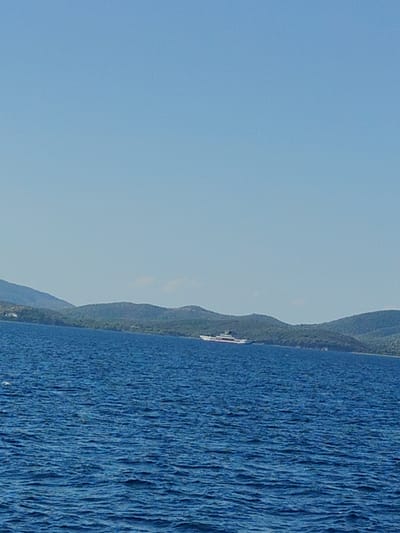 A wide shot captures a serene seascape with a large white yacht sailing on the deep blue water. In the background, lush green hills with scattered trees form a scenic coastline. The sky is a clear, bright blue, suggesting a sunny day. The scene appears to be located in the Municipal Unit of Pelasgia, Greece, known for its picturesque coastal landscapes.