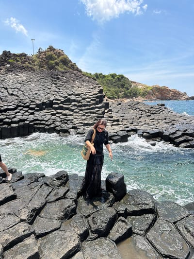 A woman stands on a rocky shoreline near Phú Lương, Vietnam. The rocks are hexagonal basalt columns, a geological formation. She is wearing a long black dress and sunglasses. She carries a woven bag over her shoulder. The ocean water is turquoise and wavy, with some splashing onto the rocks. In the background, there are more hexagonal rock formations leading up to a vegetated hillside. The sky is blue with a few white clouds.