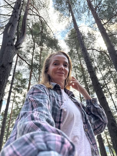 In Portovoe, Russia, a woman with blonde hair styled in a bun poses for a selfie outdoors. She is wearing a pink and grey plaid shirt over a white t-shirt, and she is touching her hair with her right hand. The background consists of tall pine trees reaching towards a bright, cloudy sky. The low-angle perspective emphasizes the height of the trees.