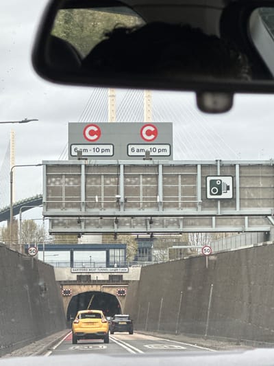 The image shows a view from a car driving on a road leading to the Dartford West Tunnel in Dartford, United Kingdom. The tunnel entrance is visible, with a yellow car approaching it. A sign indicates that the tunnel is one mile long. Road signs specify a speed limit of 50 and charging zone hours between 6 AM and 10 PM. A camera icon is present. A bridge structure is partially visible to the side of the road, with a car mirror reflecting a green landscape.