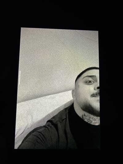 The image shows a black and white photograph of a man's face. His head is tilted, and he is wearing a dark garment. He has a tattoo on his neck. The background is a grainy gray with some white areas. The image is displayed on a screen, likely a tablet or phone, with a black border. The screen appears to have some cracks. There is a white bar at the bottom of the screen. The photograph does not depict any identifiable location in Kalitinovo, Bulgaria.
