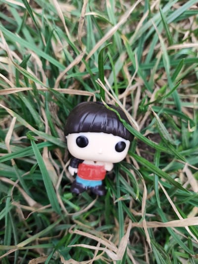 A Funko Pop figurine, resembling a child with a dark brown bowl cut hairstyle, is positioned in a patch of grass. The figure has pale skin, large black eyes, and a small mouth. It wears a red shirt with a letter "T" design and blue shorts. The grass is a mix of green and brown, suggesting a lawn or field. The figurine is the primary focus, centered within the frame of the shot. The photograph appears to be taken outdoors, in daylight. The location in the city of Yerzhovo, Moldova, is not discernable from the image alone.