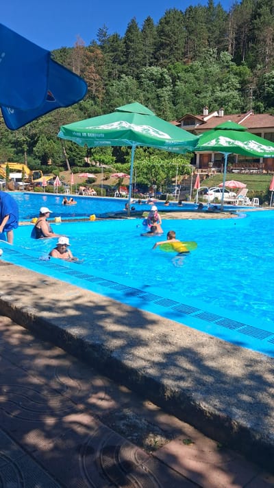 A bright, sunny day at an outdoor swimming pool in Marchevo, Bulgaria. The pool, filled with clear blue water, is the central focus of the image. Several people are enjoying the water, including adults and children. Some are swimming, while others are relaxing near the edge. One child is wearing a yellow inflatable ring. In the background, a lush green forest rises up a hillside, dotted with buildings. Large green umbrellas provide shade over parts of the pool area. A yellow excavator is visible near the pool's edge, suggesting recent construction or maintenance work. The scene conveys a sense of leisure and enjoyment on a warm day.