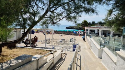 The image depicts a sunny day at a beachside establishment in Leporano, Italy. In the foreground, a concrete path with white railings leads towards the beach. To the right, a modern structure with glass railings and outdoor seating suggests a cafe or restaurant. On the left, a white building, possibly a changing room or service area, is visible under the shade of a large tree.
The beach itself is sandy, dotted with numerous green umbrellas and sunbathers. The turquoise sea stretches towards the horizon, with a rocky coastline visible in the distance. Several people are scattered across the beach and seated at tables outside the establishment. On the beach, a group of three young men are seated at a table. A motorcycle is parked near the entrance of the establishment. The overall scene conveys a relaxed atmosphere of a summer day at the seaside.