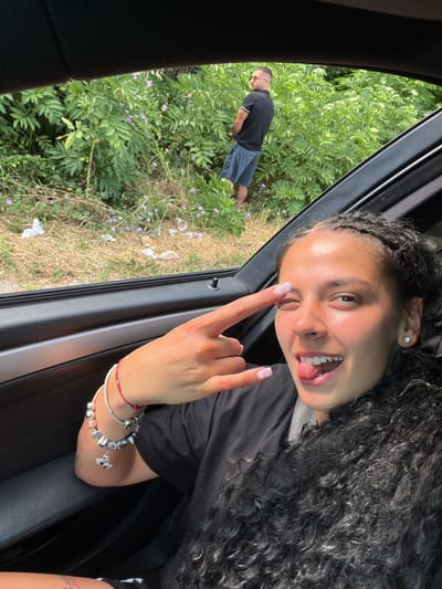 This media file features two individuals. In the foreground, a young woman with long, dark, curly hair and braids is leaning out of a car window. She is smiling broadly, with her tongue sticking out and one eye winking. She is wearing a black t-shirt and has multiple bracelets on her right wrist, including a charm bracelet. Her fingernails are painted with a light pink and white heart design. She is making a "rock on" hand gesture.
In the background, a man is visible standing outdoors amidst lush green foliage and dry grass. He is wearing a black polo shirt and grey shorts, with sunglasses on his head. He appears to be urinating. The scene suggests a casual, possibly roadside, setting. Small purple flowers are scattered throughout the greenery. Some litter, including plastic cups and wrappers, is visible on the ground near the man.
The image is taken from inside the car, looking out the open window. The lighting indicates it is daytime, likely sunny. The overall mood is playful and carefree, as conveyed by the woman's expression and gesture. The text "Garnia, Bulgaria" is provided as location context.