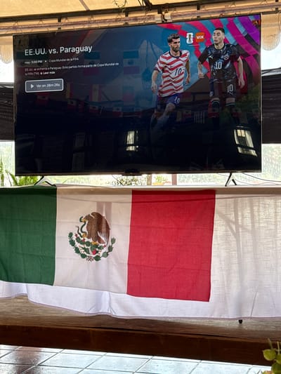 The image shows a television screen displaying a soccer match between EE.UU. (USA) and Paraguay, with the FIFA World Cup logo visible. Text on the screen indicates the match is airing live at 5:00 PM and is part of the FIFA World Cup. Below the television, a Mexican flag is draped over a surface, possibly a table or railing, with its distinctive eagle emblem at the center. The background of the image reveals a bright, possibly outdoor setting with foliage and windows. The scene is likely set in Tepeji de Ocampo, Mexico, given the presence of the Mexican flag, which is often displayed in public and private spaces in the country, especially during national or sporting events. The television screen displays information in Spanish, suggesting a Spanish-speaking region.