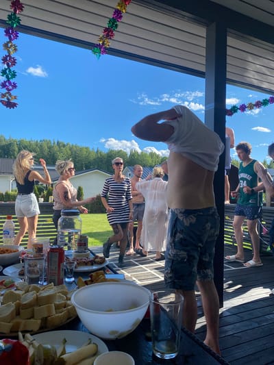 This is a photograph taken outdoors on a sunny day, likely in Joensuu, Finland, given the context. The scene appears to be a casual gathering on a wooden deck or patio, possibly a backyard barbecue or a social event.
Several people are visible, dressed in casual summer clothing. In the foreground, a person's torso is prominent, with their white t-shirt pulled up, revealing their stomach and floral-patterned shorts. Other individuals are standing and interacting, including a man in a green basketball jersey and patterned shorts, and a group of women. One woman is wearing a black and white striped dress, another a striped tank top and white shorts, and a third has blonde hair and is wearing a black top and white shorts. The overall mood seems relaxed and cheerful.
On a table in the foreground, various objects are laid out, including a large white bowl, plates of food like sliced bread and what appears to be some sort of pastry or cake, glasses, and cans of beverages. One can has visible text, "Le Coq". Another plastic bottle has the brand name "elle". Colorful flower-shaped garlands are strung along the roofline of the structure, adding a festive touch. The bright blue sky with scattered white clouds suggests it is daytime, with good weather. The presence of trees and a house in the background further establishes the outdoor, residential setting.