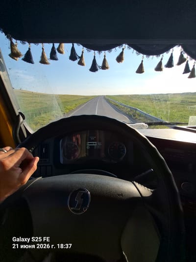 The image captures a first-person perspective from the driver's seat of a truck, looking through the windshield. A steering wheel with a logo is prominently in the foreground. The dashboard displays some gauges and digital readouts. Hanging from the sun visor above the windshield are several decorative tassels, alternating in color between yellow and dark blue. Outside, a paved road stretches into the distance, flanked by vast, green, rolling hills under a bright blue sky with scattered clouds. The environment appears to be rural and open. Text overlaid on the bottom left corner of the image indicates the device used was a "Galaxy S25 FE" and the date and time are "21 июня 2026 г. 18:37". The mention of Златополье, Kazakhstan, suggests this is the geographic context of the scene.