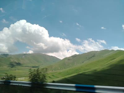This is a horizontal, eye-level, medium shot of a vast, rolling landscape under a bright blue sky filled with large, white cumulus clouds. The scene appears to be a rural area, possibly on the outskirts of Tsilkar, Armenia, given the general characteristics of the Armenian countryside.
The foreground is dominated by a metal guardrail with blue and white accents, suggesting the viewpoint is from a vehicle or a roadside. Blurry green foliage is visible just behind the guardrail, indicating movement or a shallow depth of field.
The midground and background feature lush, green, undulating hills and mountains. The sunlight casts distinct shadows across the slopes, creating a dynamic interplay of light and dark. Patches of darker green and some lighter, possibly cultivated fields, are visible in the valleys between the hills. Sparse trees dot some of the higher inclines. The overall impression is one of natural beauty and expansive open space. There are no people or animals visible in the image. The weather appears to be pleasant, with clear skies and fair-weather clouds.