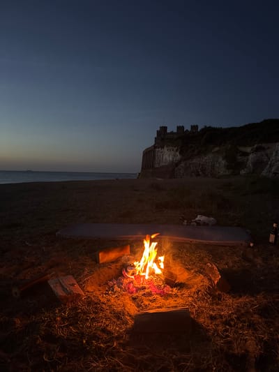 A campfire burns on a beach in Thanet, United Kingdom, at dusk. In the background, a castle sits atop a cliff overlooking the sea. The scene is peaceful and serene, with the flames of the fire casting a warm glow on the surrounding sand and debris. A camping mat and some bottles are placed nearby. A ship is visible on the horizon. The sky is a deep blue, with a few stars starting to appear.