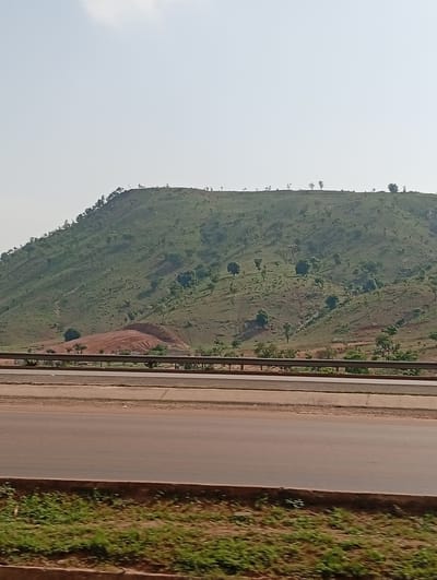The image shows a hill with green vegetation and scattered trees, possibly in Kugbo, Nigeria, as indicated in the prompt. A section of exposed reddish-brown soil is visible on the hillside. A highway with a metal guardrail runs in the foreground. The sky is overcast with a light haze. A strip of green grass grows along the highway.