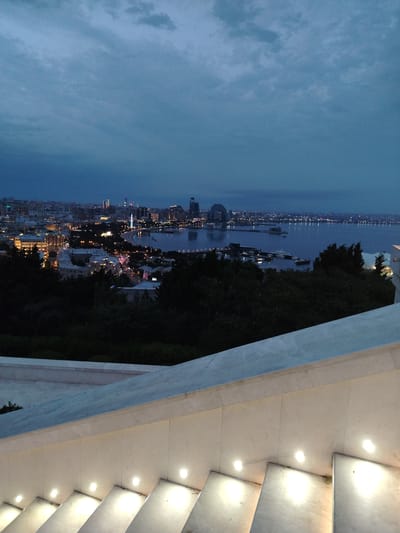 The image captures a twilight view of the Baku skyline in Sabail Raion, Azerbaijan, taken from an elevated position. In the foreground, a modern marble staircase with integrated lighting curves upwards, leading the viewer's eye towards the cityscape. The city lights are illuminated, reflecting on the calm waters of the Caspian Sea. Various buildings, including distinctive modern structures like the Flame Towers and the Crescent Development, are visible along the waterfront and further inland. Trees are scattered throughout the middle ground, separating the staircase from the dense urban landscape. The sky is a deep, dusky blue with scattered clouds, indicating the transition from day to night. Boats and ships are docked or anchored in the bay. The overall scene is serene and depicts a vibrant coastal city at dusk.