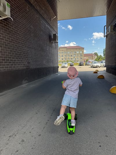 A rear view of a young child wearing a pink hat and blue denim shorts rides a green scooter. The child is in a paved walkway between two brick buildings. In the background, cars are parked in front of an apartment building under a blue sky with scattered clouds. The location is in the city of Vyazniki, Russia.