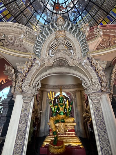 The image depicts an ornate interior, potentially a temple or religious site in Samrong Nuea Subdistrict Municipality, Thailand. A green and gold statue stands within a decorated alcove. Elaborate carvings and mosaics adorn the walls and ceiling, featuring intricate patterns and designs. A stained-glass dome-like structure appears in the upper left portion of the frame. The statue is a figure in either gold or bright green color, and is sitting on an elevated platform also in gold. It is inside what appears to be an archway and a room that is ornately designed with carvings.
