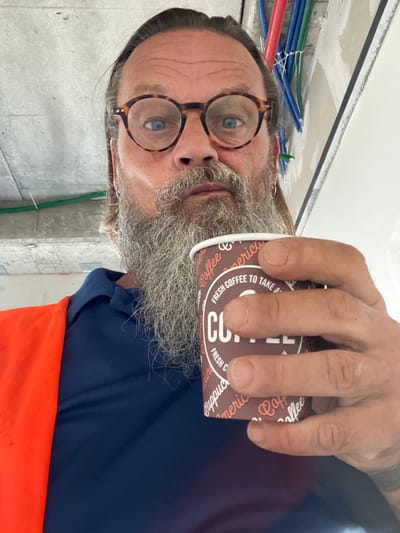 In Ramot HaShavim, Israel, a man with a long beard and round, tortoiseshell glasses is taking a selfie. He is wearing a dark blue collared shirt and an orange high-visibility vest. The man holds a brown paper cup with the word "COFFEE" printed in large white letters, along with text that reads "FRESH COFFEE TO TAKE A..." and other partial words. Behind him, the environment appears to be an indoor construction site, indicated by exposed concrete ceilings, wiring, and conduit pipes.