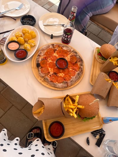 An overhead, close-up shot shows a table laden with food and drinks, likely at a cafe in Ptghni, Armenia. The table has a white marble top with grey veining. In the foreground, a white and black polka dot dress is visible, along with black sandals with gold buckles. A person's feet wearing these sandals are positioned near the bottom of the frame.
The food spread includes a pepperoni pizza on a wooden circular board, several servings of french fries in brown paper cones, and two burgers on wooden trays. One burger is complete with bun, lettuce, and tomato. Another burger bun is visible in the lower right portion of the image. Small bowls of ketchup are scattered amongst the fries. A white oval dish holds what appear to be fried cheese balls, accompanied by two small bowls, one with a pinkish sauce and the other with ketchup. A glass bottle of "MAX & JACK'S" cherry-flavored drink sits to the left of the pizza. Other objects on the table include white plates, a fork, an ashtray with cigarette butts, a lighter, a bottle cap, and a clear plastic bottle of water. The background shows hints of outdoor seating with tiled flooring and a blurred figure seated at a table.