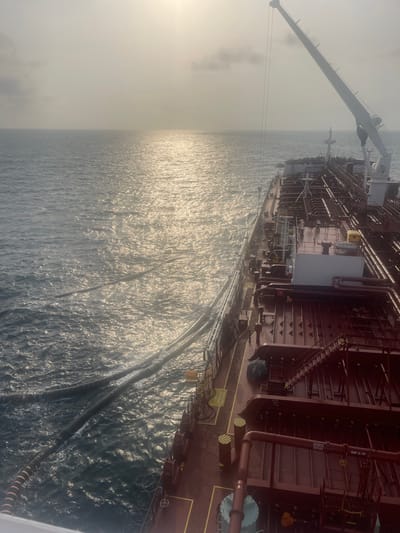 A large vessel with intricate piping systems and a prominent crane sits on the water's surface. Hoses extend from the side of the ship into the ocean, creating ripples on the water. The sky is hazy, and the sun reflects off the water. The overall scene depicts a ship engaged in activity on the open sea.