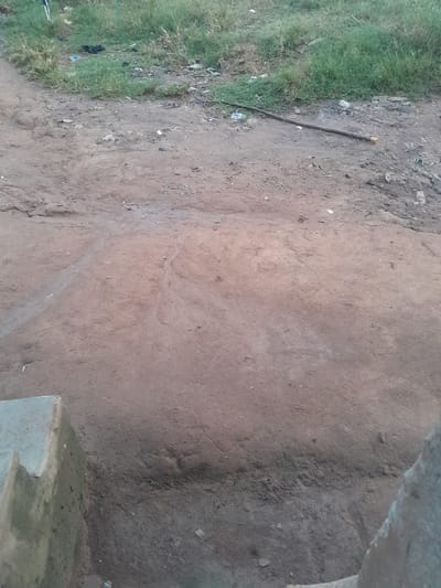 This image depicts an outdoor ground-level scene in Ifo, Nigeria. The foreground shows reddish-brown compacted dirt ground, uneven and marked with subtle depressions and faint paths. Partial views of two worn, light-grey concrete structures are visible at the bottom left and right edges, likely foundation elements or steps. In the midground, a dark brown stick approximately 0.75 meters long, with a lighter tip on its right end, lies horizontally on the dirt. The stick is positioned roughly parallel to the bottom edge of the image, slightly angled upwards to the right. The background features a strip of unkempt green grass and weeds bordering the dirt, extending across the upper portion of the frame. This grassy area and the adjacent dirt contain scattered litter, including crumpled plastic bags, white wrappers, and various unidentifiable small debris. No people, animals, or active interactions are visible. The lighting suggests daylight conditions, casting soft illumination across the scene.