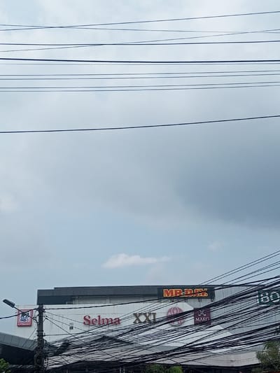 An exterior shot shows a multi-tenant building under a cloudy sky, obscured by numerous power lines. Prominent signage includes "MR.DIY," "Selma," and "XXI." A smaller sign reads "MAKO." "INDO" is also visible on another sign. The building appears to be a retail complex. A streetlight is present, along with green foliage at the bottom of the frame.