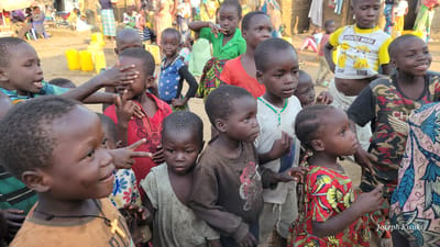 In the city of Bunia, Democratic Republic of the Congo, a group of approximately 15 children are gathered outdoors. The children, ranging in age from toddlers to young teenagers, are primarily of African descent. They are dressed in casual, brightly colored clothing, including t-shirts, shirts, and patterned dresses.
In the foreground, a young boy in an orange t-shirt with a yellow cross pendant is looking to his right. To his left, a younger girl in a red and yellow floral dress is looking to her left and pointing with her right index finger. To the right of the boy in the orange shirt, another child is wearing a camouflage t-shirt.
Behind these children, more children are visible. One boy wears a white t-shirt with a yellow and blue Pikachu design and the words "CURRENT MOOD" printed on it. Another child in a green shirt is seen in the background. The children appear to be interacting with each other, with some looking towards the camera and others engaged in conversation or play.
The environment is somewhat dusty and appears to be a public space, possibly a market or a gathering area, with yellow jerry cans visible in the middle ground. The lighting suggests it is daytime, with sunlight casting shadows. The overall scene depicts a lively gathering of children in a community setting.
