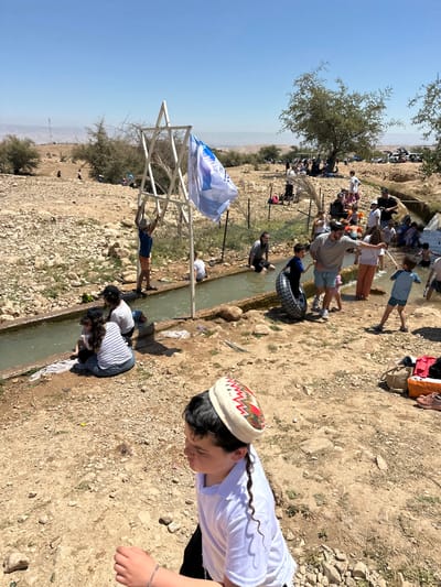 The image captures a scene outdoors in what appears to be a rural or semi-arid environment with rocky terrain. A narrow irrigation channel runs through the landscape, attracting a group of people engaging in various activities. Some individuals are partially submerged in the water, possibly washing items or cooling off. A Star of David structure adorned with an Israeli flag is visible nearby. The location is likely in Arvot Hayarden Regional Council, Palestinian Territory, given the information provided. A young boy wearing a kippah is in the foreground.