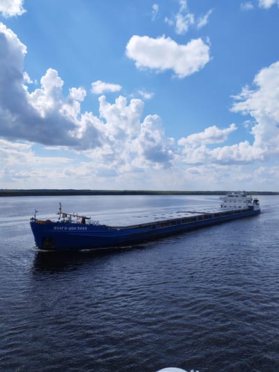 A blue cargo ship labeled "ВОЛГО-ДОН 5068" is navigating a wide river under a partly cloudy sky. The distant shore is lined with trees. The location, though unspecified in the prompt, would be on the Volga-Don Canal, as the ship's name indicates. The presence of the Volga-Don Canal in the image suggests it may be close to Pruzhinino, Russia.