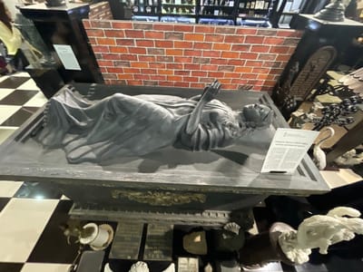 The image depicts a museum exhibit featuring a sculpture of a reclining figure, possibly a representation of death or a saint, lying in a sarcophagus-like structure. The sculpture is draped in flowing fabric, and the figure's hands are clasped together in prayer. Behind the sculpture is a brick wall backdrop. The scene is set in what appears to be a dimly lit museum with a checkered floor. Several other exhibits and decorative elements are visible in the background and foreground, including small figurines and ornamental pieces. A plaque with text is placed next to the sculpture, providing information about the artwork and artist, identified as "Rafal Monti (1818-1882), master of monumental sculpture." The text on the plaque is in Russian. There are no people clearly visible interacting with the exhibit. The image is a wide shot, capturing the entire display. The overall atmosphere is somber and artistic, typical of a museum setting. The location is confirmed as the Museum of History and Local Lore in Voskhod, Russia, based on the information on the plaque.
