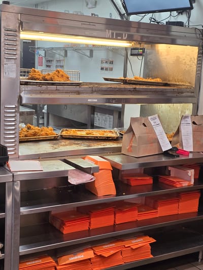 This media file captures a close-up, low-angle view of a fast-food restaurant's food preparation and packaging area, likely Popeyes, given the prominent branding on the orange bags and packaging. The scene is dominated by stainless steel surfaces and a bright overhead fluorescent light, suggesting an indoor, commercial kitchen environment.

The primary subjects are food and packaging supplies. In the foreground, trays of golden-brown, fried chicken are visible under heat lamps, indicating the food is being kept warm for service. To the right, stacks of bright orange paper bags, many bearing the "Popeyes" logo, are neatly organized in compartments, ready for customer orders. Some of these bags appear to contain receipts or order slips. A small digital control panel with red and green buttons is also visible, likely part of the cooking or warming equipment.

The overall impression is one of a busy service environment. The presence of prepared food ready for distribution and an abundance of packaging materials suggests an active period of operation, possibly during a lunch or dinner rush. No people are visible in the frame. The lighting is artificial and intense, casting warm tones on the chicken and creating reflections on the metal surfaces. The text "MT-U", "CLASSIC", and "SPICY" are visible, likely labeling different food options or sections of the equipment. The location context of Alpharetta, United States, places this scene within a typical fast-food setting in America.

---
**Summary:**

*   **Subjects:** Fried chicken, orange paper bags with "Popeyes" branding, stainless steel kitchen equipment, digital control panel.
*   **Scene/Setting:** Indoor commercial kitchen of a fast-food restaurant.
*   **Activity:** Food preparation and packaging. Fried chicken is displayed under heat lamps, and packaging supplies are ready for use.
*   **Notable Details:** Prominent "Popeyes" branding, artificial lighting, stainless steel surfaces, labels "MT-U," "CLASSIC," and "SPICY."
*   **Location Cues:** "Alpharetta, United States" suggests a US-based Popeyes restaurant.