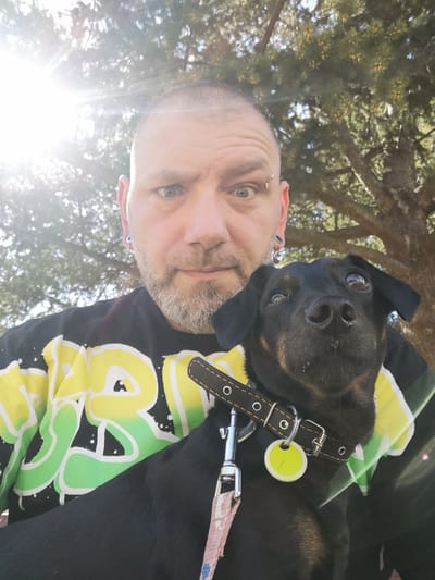 A medium shot depicts a man holding a small black dog. The man has short hair and a light-colored beard, and he is wearing a black T-shirt with yellow and green designs. He has piercings in his ears and an eyebrow piercing. The dog is wearing a black collar with a tag. The background shows a tree with dense foliage and sunlight filtering through the leaves. The location cannot be determined to be in Pardubice, Czechia, from the given information.