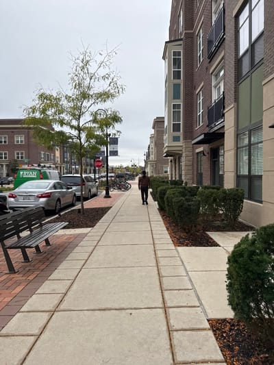 This image captures an urban street scene in South Bend, United States, specifically on Green Street, indicated by signage on a park bench. The setting is a cloudy day, characterized by an overcast, light gray sky.
In the foreground, a black metal park bench is positioned on a red brick paver section of the sidewalk on the left. The bench's backrest has "GREEN STREET" and "SOUTH BEND" carved into it. To the right of the bench, the sidewalk transitions to light gray concrete, extending into the background.
A single individual, wearing a dark brown long-sleeved top and dark pants, walks away from the viewer along the concrete sidewalk, heading towards the background. The person's hair appears dark, possibly tied back.
To the left of the sidewalk, a young tree with sparse green foliage and some bare branches is planted in a mulched area. Beyond the tree, a street runs parallel to the sidewalk. Parked along this street are several vehicles: a silver sedan with a partially visible license plate ("M 088"), a silver minivan or SUV, and a green utility van with "RYAN" printed in white on its side. Further down the street, multiple bicycles are parked together. A red hexagonal "STOP" sign is visible on a pole near the parked vehicles.
On the right side of the sidewalk, a series of manicured green hedges are planted in mulched beds, separating the pedestrian path from a multi-story building. This building is constructed of reddish-brown brick with light-colored stone accents on the ground floor. It features numerous windows, some with dark frames, and bay windows extending outwards. Black metal railings are visible on some balconies. Other multi-story brick buildings, consistent in style, extend into the background on both sides of the street. Black lampposts with banners, one clearly displaying "SOUTH BEND," line the street. The scene is illuminated by natural, diffused light, indicating an absence of direct sunlight and strong shadows.