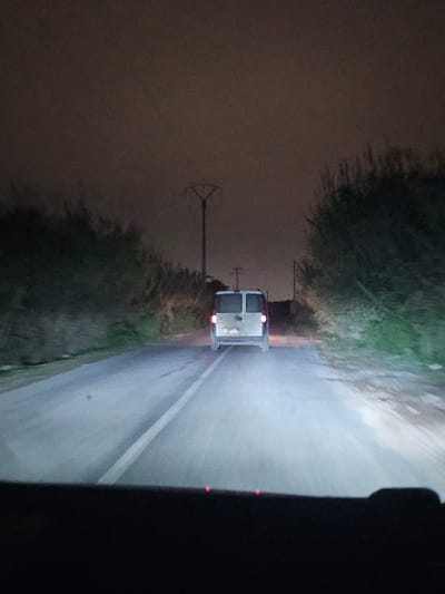 A white van travels down a dark, wet road at night. The vehicle is seen from behind, its taillights glowing red. The road has a single white line marking the center. Both sides of the road are lined with dense foliage and a utility pole is visible on the left side. The image appears to be taken from inside another vehicle following closely behind. A small portion of the dashboard is visible on the right side of the frame.