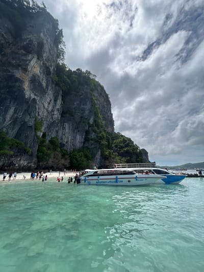 This image depicts a scenic view of a tropical beach with a steep, vegetated cliff face rising behind it. The water in the foreground is a clear turquoise, showing ripples on its surface. A white and blue speed boat, labeled "UNSEEN ANDAMAN TOUR," is anchored close to the shore. Numerous people are visible on the sandy beach and wading in the shallow water. The sky is overcast with dramatic clouds. The location is identified as Ko Phi Phi, Krabi Province, Thailand, known for its limestone karst formations and beautiful beaches.
