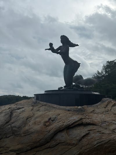 A bronze statue of a mermaid holding a child stands on a rocky outcrop in Ao Hin Khok, Thailand, under a cloudy sky. The mermaid, with flowing hair and a scaled tail, is depicted in profile, extending one arm towards the child. The child, held in the mermaid's other arm, is small and appears to be reaching out. The statue is mounted on a dark, circular base. Behind the mermaid statue, partially obscured by the mermaid's tail, is another bronze sculpture of what appears to be a fish or sea creature, with its head and fins visible. The base of the statue rests on large, weathered rocks that form the foreground of the image. In the background, lush green trees cover a hillside, suggesting a coastal or island environment. The sky is overcast, with dramatic cloud formations that create a somewhat somber atmosphere. The overall scene is static, with no visible human activity or interaction beyond the depicted statue.