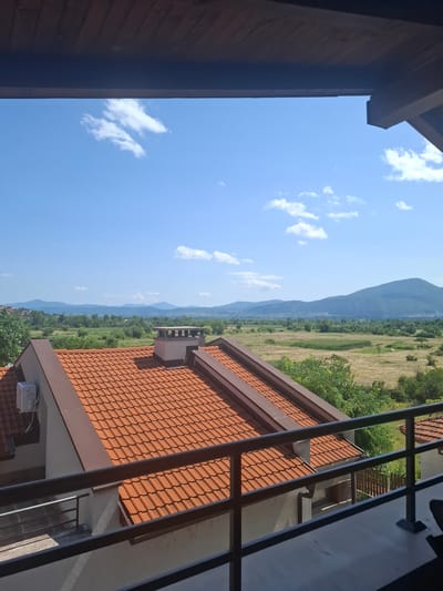 The image shows a view from a balcony overlooking a landscape in Ognyanovo, Bulgaria. The foreground features a tiled roof of a building with a chimney and an air conditioning unit. A metal railing is visible in front of the balcony. The middle ground consists of fields and trees. In the background, rolling hills and mountains under a blue sky with scattered clouds complete the scene. There are no people visible in the image.