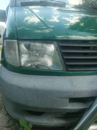 The image shows the front of a green van, likely in Velyka Dymerka, Ukraine. The focus is on the vehicle's front bumper, headlight, and grill. The bumper is a dull gray and appears to have scuffs and scratches, indicating it's seen some use. The headlight is clear with visible internal reflectors and a patterned lens. The grill is black and has a horizontal slat design.
The van's hood is a shiny green, reflecting the sky, trees, and clouds, suggesting it's daytime with partly cloudy weather. A bit of a metallic bull bar or guard is visible on the right side of the image, protruding from the lower part of the bumper. There is some green foliage in the foreground at the bottom left, adding a touch of nature to the scene. The overall impression is a mundane, everyday view of a vehicle parked outdoors. No people are visible, and no specific activity is taking place; it's a static shot of the vehicle's front. No visible text is present in the image.