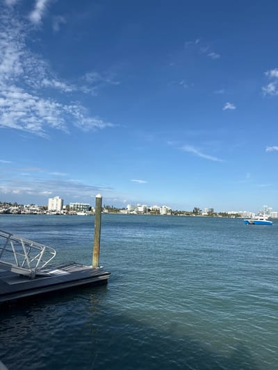 A serene view of a pier extending into calm water, with buildings lining the distant shore. A blue boat is visible on the water. The sky is predominantly blue, adorned with scattered wispy clouds. The overall scene evokes a peaceful coastal atmosphere.