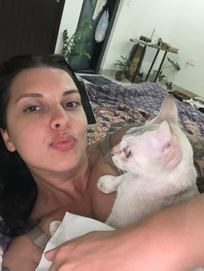 A selfie taken in Hoi An, Vietnam shows a woman with dark hair lying on a bed with a light-colored cat. The woman is looking at the camera with her lips pursed. The cat is nestled in her arms, looking away from the camera. The background shows a patterned blanket on the bed, a dark wooden door, and a shelf with plants and objects. The scene appears to be indoors, in a bedroom.